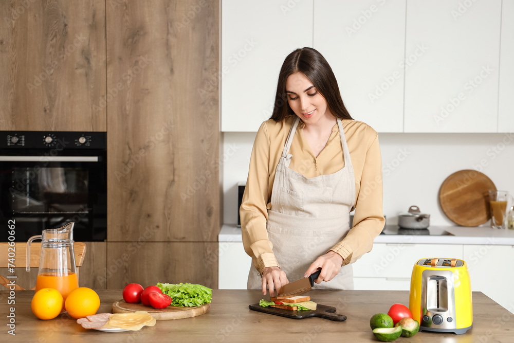 Young beautiful happy woman cutting tasty sandwich in kitchen