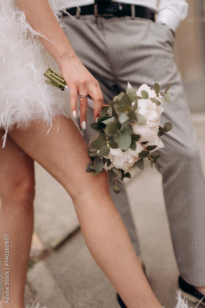 A close-up of the bride's bouquet of white peonies, roses, and eucalyptus. The bride is holding a bouquet in her hand in a white elegant short satin dress decorated with feathers A close-up of the bride's bouquet of white peonies, roses, and eucalyptus. The bride is holding a bouquet in her hand in a white elegant short satin dress decorated with feathers