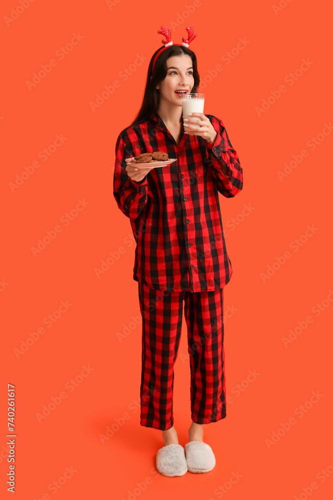 Beautiful young woman in checkered pajamas with sweet cookies and glass of milk on orange background