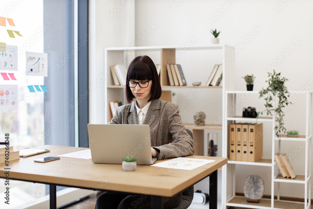Confident young employee in formal wear unlocking laptop keyboard with password at desk in modern office. Caucasian female worker investigating trademarks using search engine in creative workplace. Confident young employee in formal wear unlocking laptop keyboard with password at desk in modern office. Caucasian female worker investigating trademarks using search engine in creative workplace.
