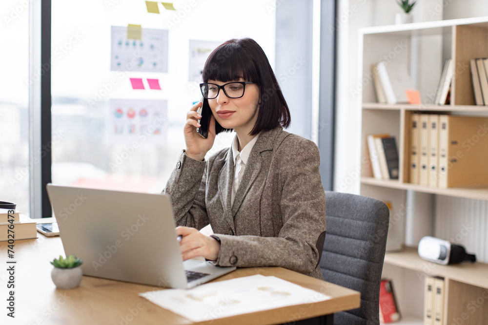 Attractive Caucasian person making important call via cell phone while scrolling web pages on laptop in workplace. Happy entrepreneur in suit negotiating startup purchase via digital devices in office Attractive Caucasian person making important call via cell phone while scrolling web pages on laptop in workplace. Happy entrepreneur in suit negotiating startup purchase via digital devices in office