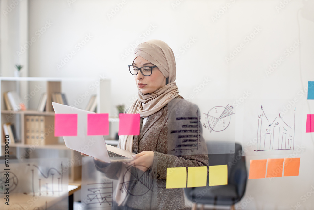 Confident muslim woman in formal wear and hijab solving business issues with laptop in hands while standing at company boardroom. Concept of modern technology and busy people. Confident muslim woman in formal wear and hijab solving business issues with laptop in hands while standing at company boardroom. Concept of modern technology and busy people.