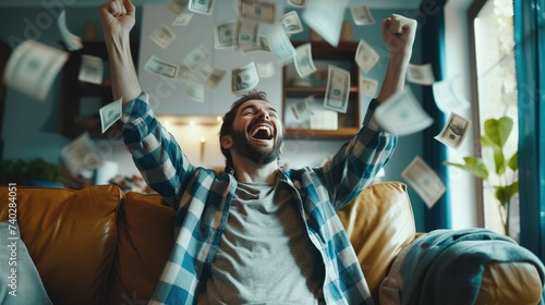 Joyful man sitting on a couch with fists raised in excitement as money cascades around him, symbolizing financial success or a lottery win. photo