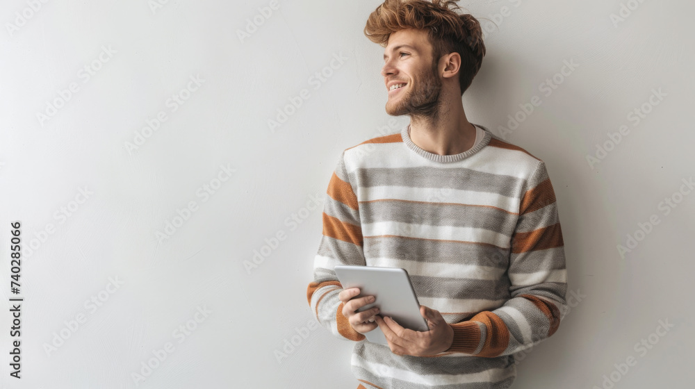 a person is casually leaning against a white wall, holding a tablet with a thoughtful smile, wearing a striped sweater.