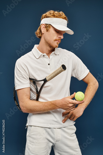 young redhead man in white clothes and sun visor holding racquet and looking at tennis ball on blue © LIGHTFIELD STUDIOS