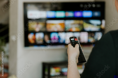 Hand holding remote control pointing to TV. Person watching smart television indoors. Blurred background. Colorful screen, copy space. Woman finger in foreground pressing button. Movie stream service