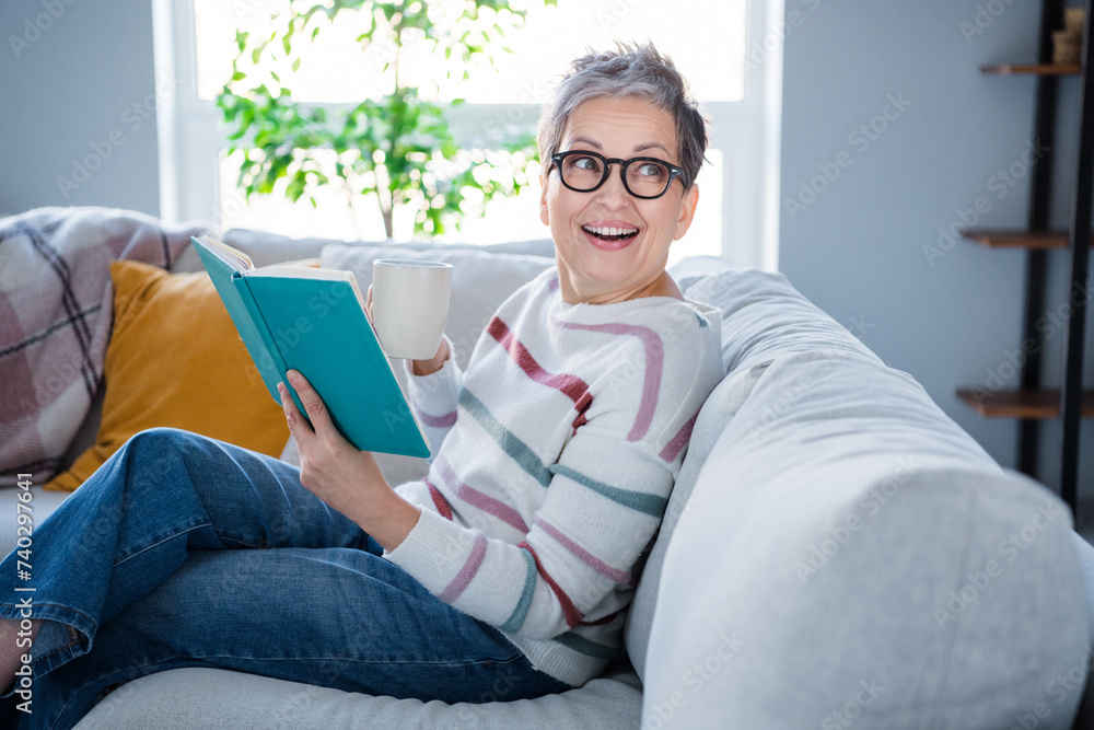 Profile portrait of positive aged person sitting sofa hold opened book coffee mug look interested away daylight flat indoors