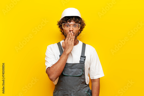 shocked Indian builder in hard hat and overalls covers his mouth with his hand on yellow isolated background, surprised Indian foreman in uniform looks at camera in amazement