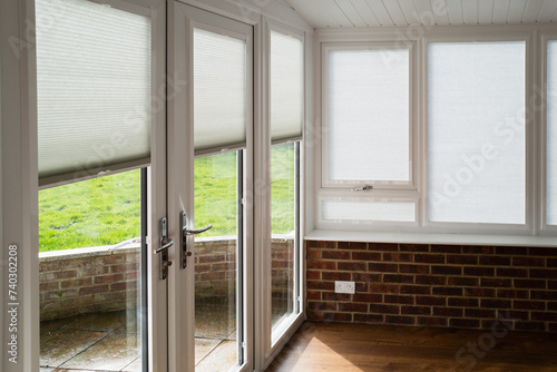 Made to measure blinds on windows and doors in a white conservatory with wooden floor and red brick walls on a sunny day. The roof is an insulated warm roof with mock beams.