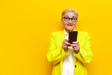 pensive old businesswoman in formal wear using smartphone on yellow isolated background, elderly pensioner grandmother in blazer holding mobile dreaming and imagining