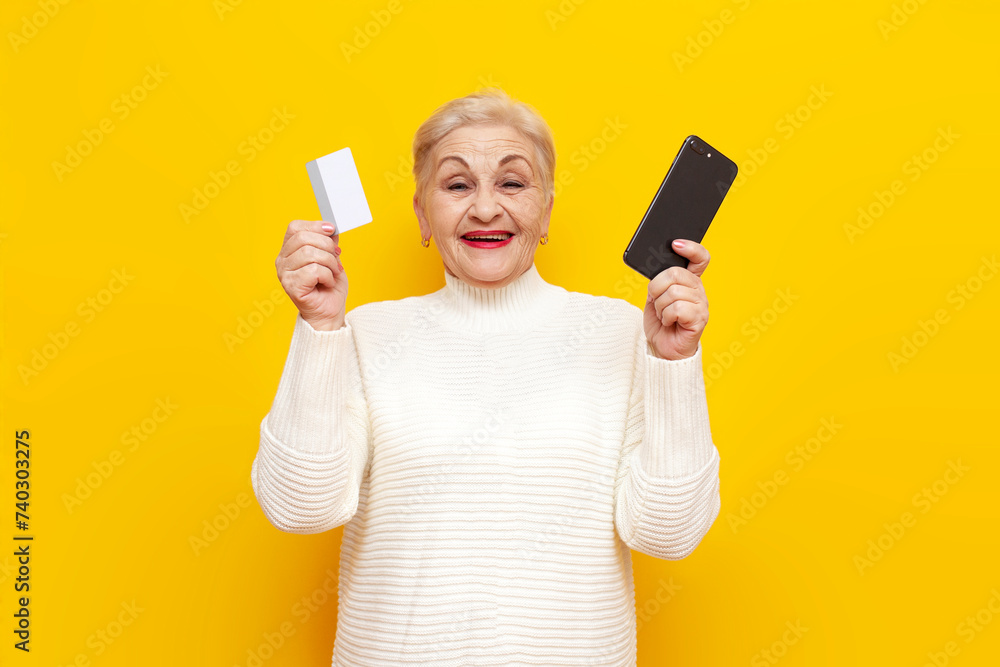 old grandmother in white sweater shows smartphone and bank card and buys online on yellow isolated background, an elderly pensioner woman holds a mobile and a credit card and smiles old grandmother in white sweater shows smartphone and bank card and buys online on yellow isolated background, an elderly pensioner woman holds a mobile and a credit card and smiles