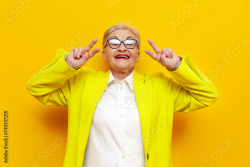 crazy old businesswoman in glasses and formal wear showing peace gesture and tongue out on yellow isolated background, elderly pensioner grandmother in blazer showing number four with fingers