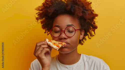 A joyful person is enjoying a slice of pizza  smiling broadly with red glasses  against a yellow background.