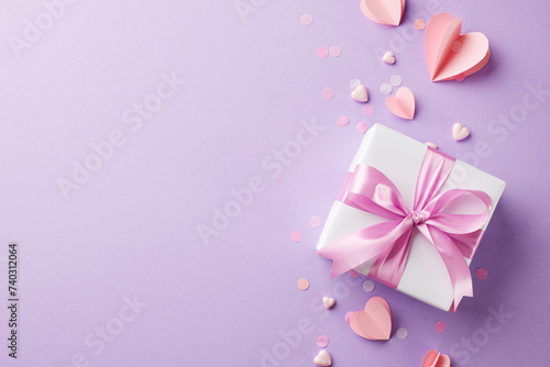 Graceful offerings: the art of giving to women. Top view shot of white gift box, pink satin ribbon, paper hearts, confetti on lavender background with space for seasonal greetings or promo content © Goncharuk film