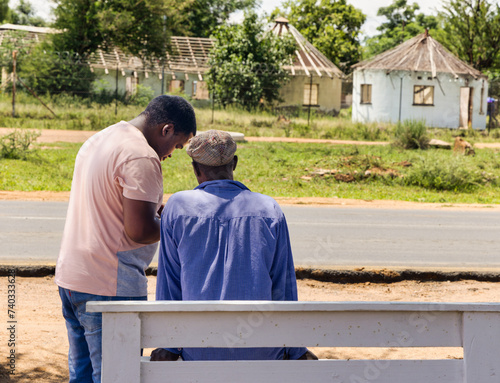 african men sited on a bench next to the road talking with his friend  in the township  rondavel in the background