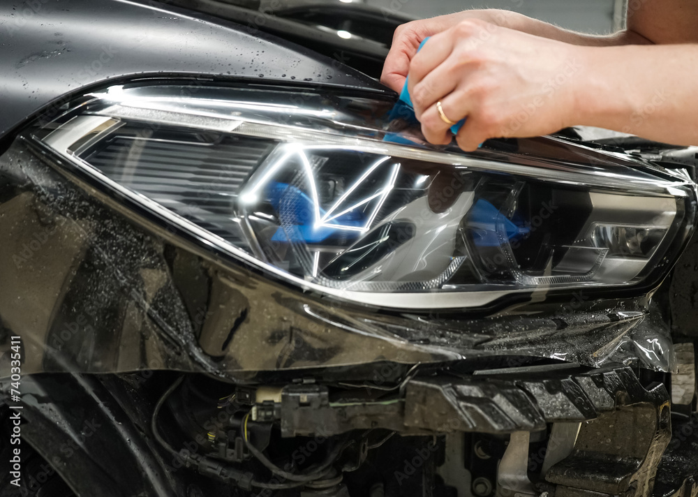 The process of installing a protective polyurethane film for paint on the front headlight of a car. Close up of paint protection film installation on headlight of modern luxury car. 
