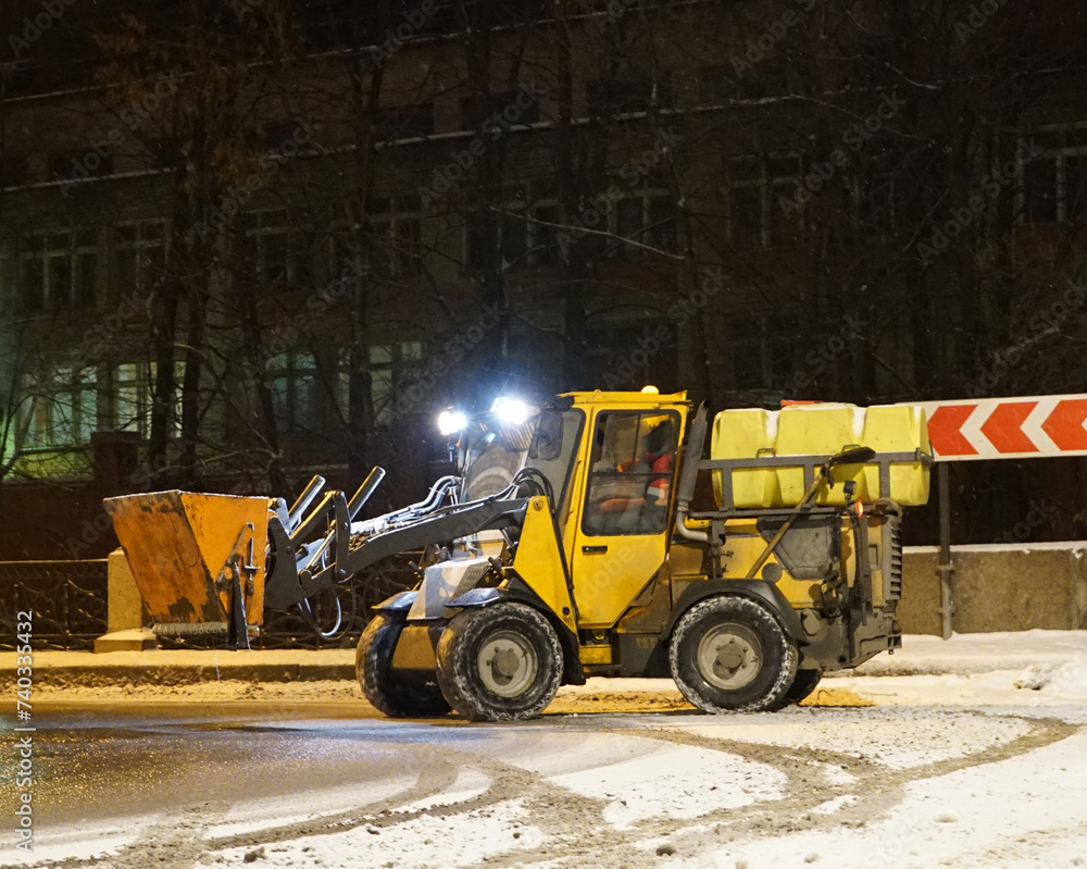 A snowplow cleans the sidewalks. A snowplow clears the street of snow at night. Selective focus. Russian winter. Snow removal.	