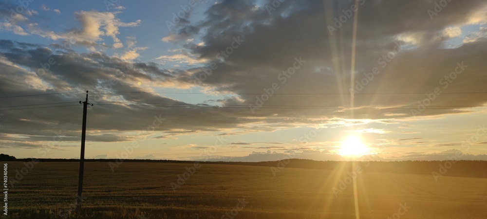 Green field at sunset. On a summer evening, small cumulus clouds hang in the blue sky. Below them is a green field with tall grass and wildflowers. In the distance the sun is setting below the horizon