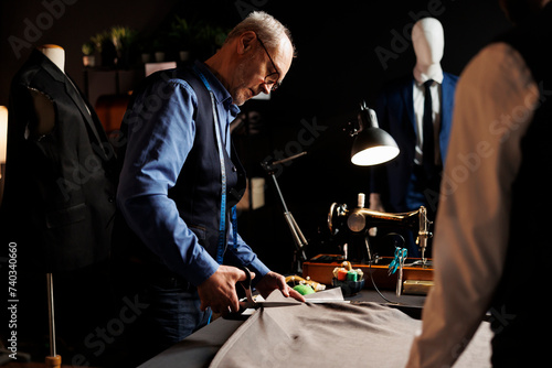 Precise experienced elderly suitmaker manufacturing suit in tailoring studio workspace, cutting fabric material with scissors. Process of manufacturing upcoming bespoke fashion design collection photo