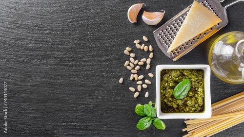 Homemade pesto sauce in small white square jar and ingredients for pasta on black slate background with copy space, top view, flat lay. Traditional Italian cuisine, recipe, restaurant menu