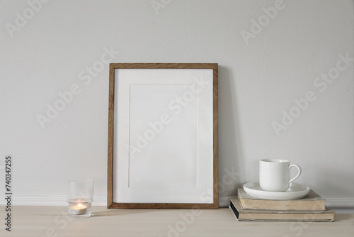 Blank vertical wooden picture frame mock up on wooden table. Modern interior. Cup of coffee on pile of vintage books, burning candle. White wall background. Scandinavian home. Poster display template.