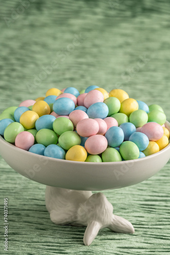 Decorative white bowl held up by the Easter Bunny full of candy coated pastel easter eggs on a textured green velvet background, Happy Easter
