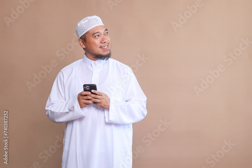 Asian Muslim man smiling when using his mobile phone while looking to the side