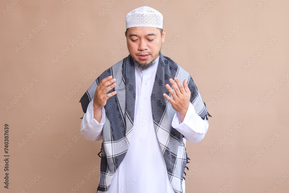 Religious Asian muslim man looking down while praying with opened palms on beige background Religious Asian muslim man looking down while praying with opened palms on beige background