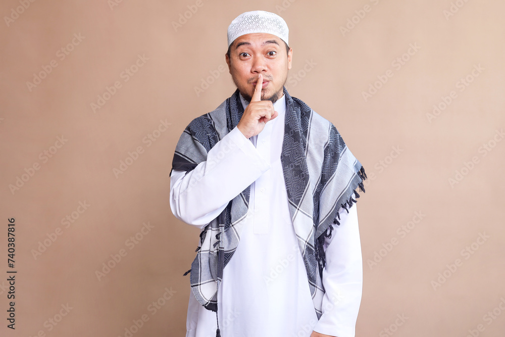 Asian Muslim man showing silence gesture with finger isolated over beige background Asian Muslim man showing silence gesture with finger isolated over beige background