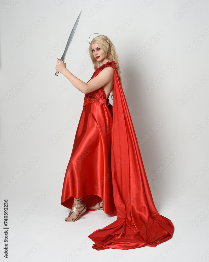 Full length portrait of beautiful blonde model dressed as ancient mythological fantasy goddess in flowing red silk toga gown, crown. walking pose, holding a sword weapon, isolated studio background Full length portrait of beautiful blonde model dressed as ancient mythological fantasy goddess in flowing red silk toga gown, crown. walking pose, holding a sword weapon, isolated studio background