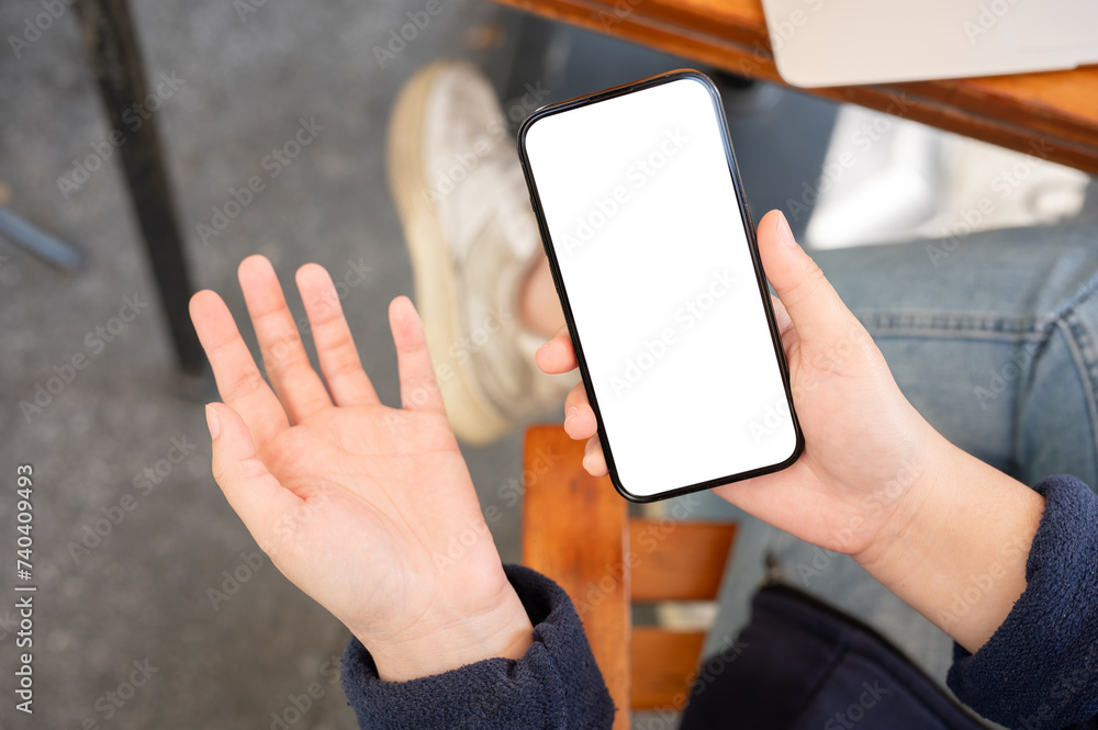 A woman showing her palm and holding a white-screen smartphone mockup while sitting in a cafe. A woman showing her palm and holding a white-screen smartphone mockup while sitting in a cafe.