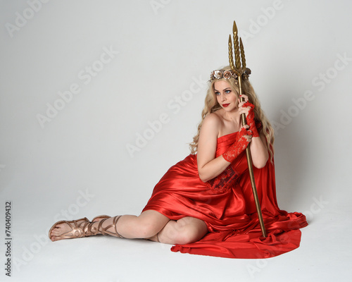 Full length portrait of beautiful blonde model dressed as ancient mythological fantasy goddess in flowing red silk toga gown  crown. kneeling pose   golden trident weapon  isolated studio background