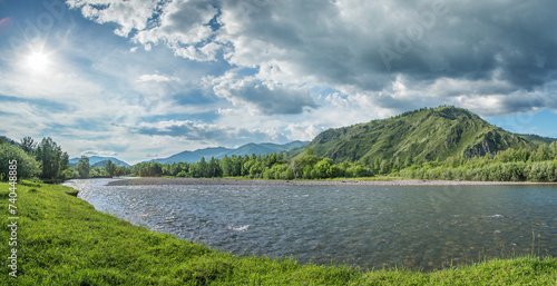 Mountain river flows among forested slopes, summer greens and shining sun