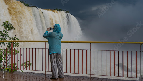 A man stands on the observation deck at the waterfall, taking pictures on a smartphone. Streams collapse from the cliff. The clothes were soaked through from the splashes. Drops on the railing.  photo