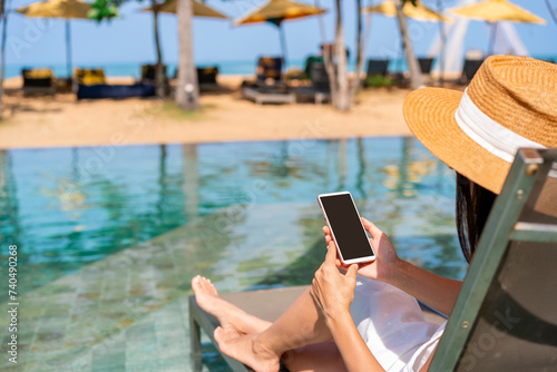Young woman traveler relaxing and using a mobile phone by a resort pool while traveling for summer vacation, Travel lifestyle concept