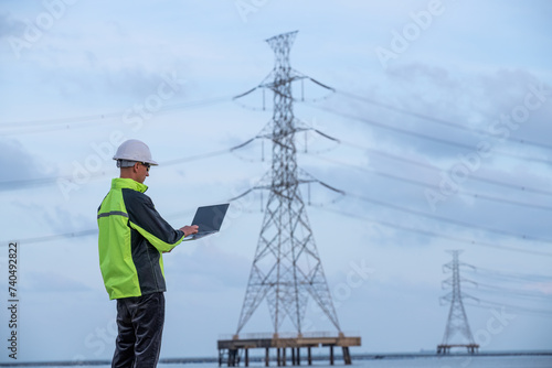 Engineer factory electrical using a notebook computer standing at a power station planning work producing electrical energy with pole high voltage electrode background
