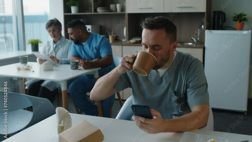 Young male doctor in medical uniform sitting in break room, drinking coffee and scrolling through social media on mobile phone during lunchtime Young male doctor in medical uniform sitting in break room, drinking coffee and scrolling through social media on mobile phone during lunchtime