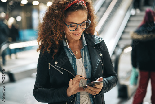 Young woman with curly hair and glasses standing in a metro station reading text messages photo