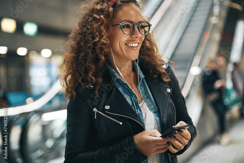 Smiling woman with curly hair and glasses standing in a metro station listening to music photo