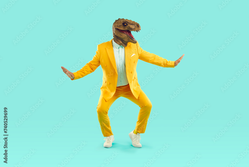 Funny showman in yellow suit and rubber dinosaur mask dancing on turquoise background. He spreads his arms to the sides and squats slightly. Banner for advertisement, marketing with funky man.