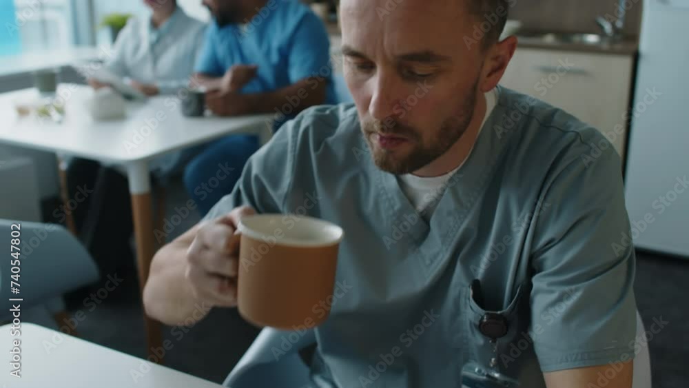 Tilt up shot of young male hospital worker wearing scrubs drinking tea and swiping on mobile phone while having lunch in break room Tilt up shot of young male hospital worker wearing scrubs drinking tea and swiping on mobile phone while having lunch in break room