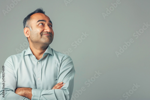 A smiling Indian businessman in a light blue shirt, arms crossed, looking away thoughtfully. © Natalya