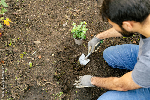 Young bearded brunette man is kneeling in the soil, planting a shrub as groundcover in the landscape. Sharing his love for gardening, he uses his thumb to gently adapt the plant into the ground