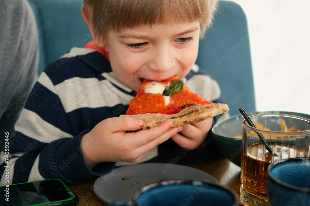 Cute healthy preschool kid boy eats a piece of cheese pizza for lunch. Charming child biting off big slice of fresh made pizza at the table in a restaurant indoor. Funny little toddler. Unhealthy food