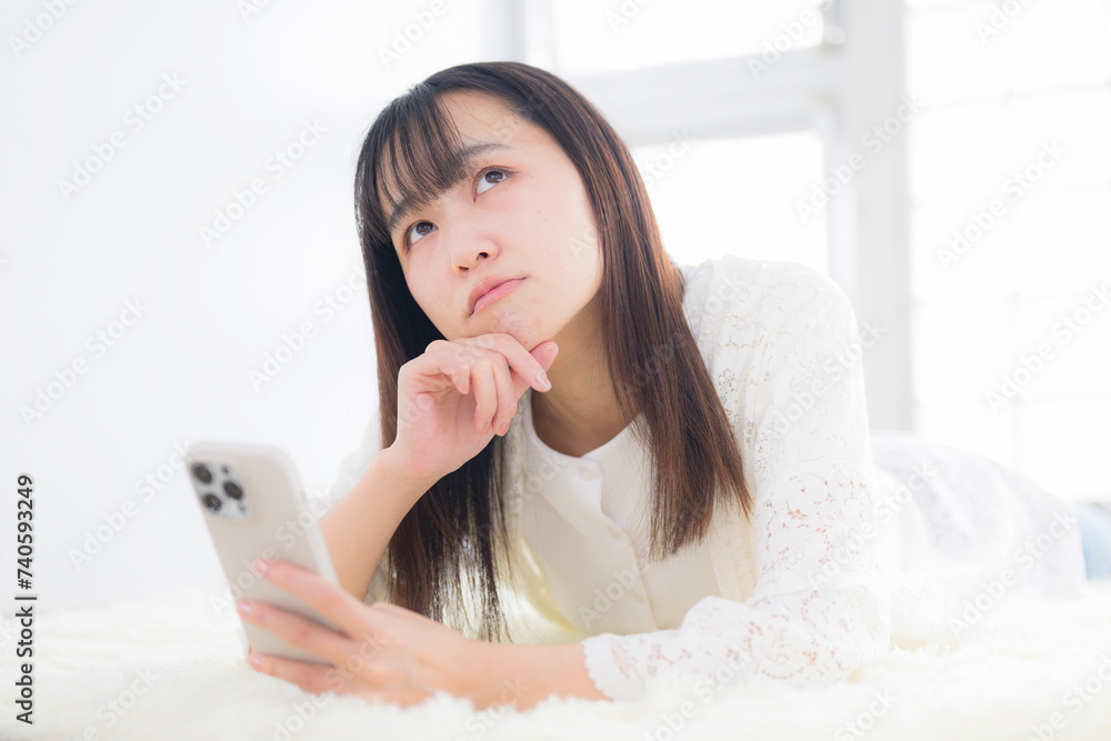 Woman lying in her room, operating her smartphone, worrying, thinking, and having trouble. Woman lying in her room, operating her smartphone, worrying, thinking, and having trouble.