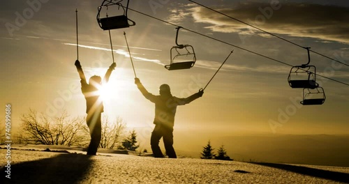 Silhouette of two friends against sunset sky raising their arms in celebration beneath a row of empty ski lift chairs. Skiers and chairlifts in winter resort. 50 fps on cinema camera photo