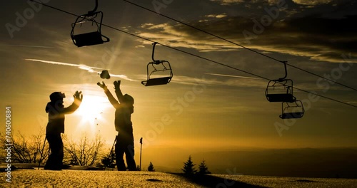 Silhouette of two friends against sunset sky throwing show in the air beneath a row of empty ski lift chairs. Skiers and chairlifts in winter resort. 120fps on cinema camera photo