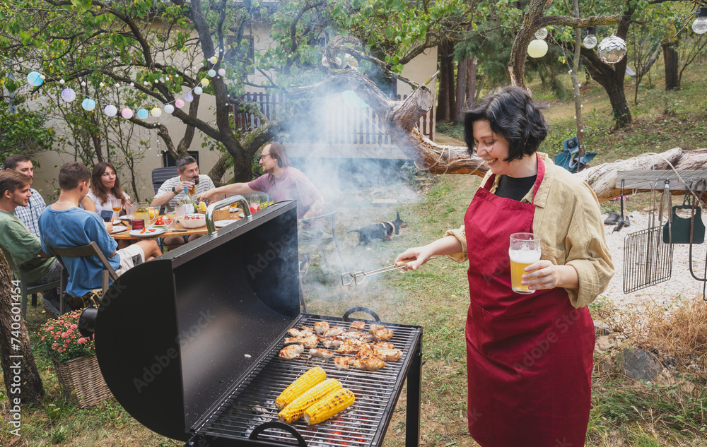 Young happy woman in apron grilling barbecue in garden for friends at country house party