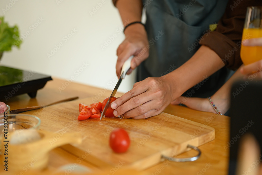 Obraz premium Young man cutting tomato on wooden board at kitchen counter. Closeup view