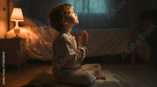 Little toddler boy kneeling on the floor of his bedroom interior late at night or in the evening, closed eyes, praying to God with his hands clasped together indoors. Asking for protection,forgiveness photo